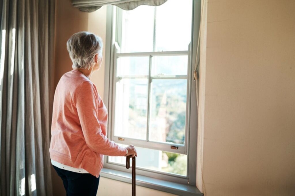 An elderly woman with a cane looks out a window