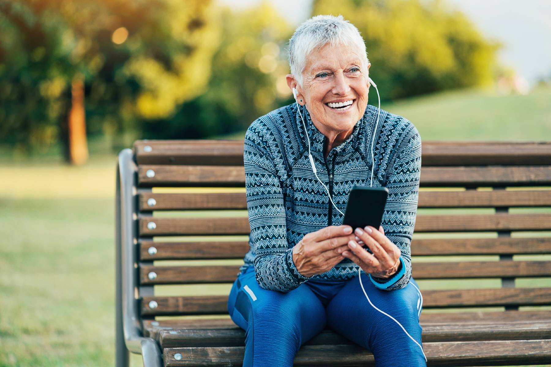 A senior woman in athletic gear stops her run and sits on a park bench to list to music on her phone