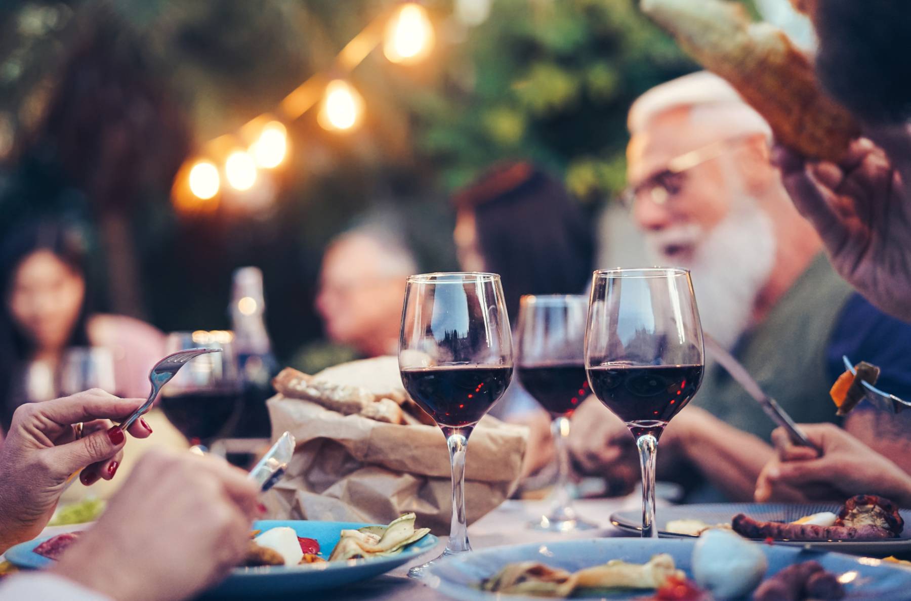 A group of people eat outside under string lights and drink red wine and eat a meal together